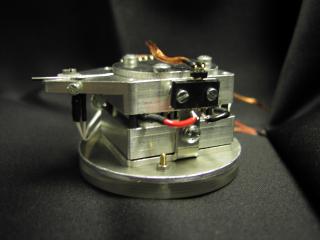 Miniatursteuerroboter mit Verdampfungsanlage, die zur Bereitstellung goldhaltiger Substanzen in der Vakuumkammer eines Rasterelektronenmikroskops (REM) dient. Der Roboter ist als "Scheibe" sichtbar und hat einen Durchmesser von ca. 5 cm.